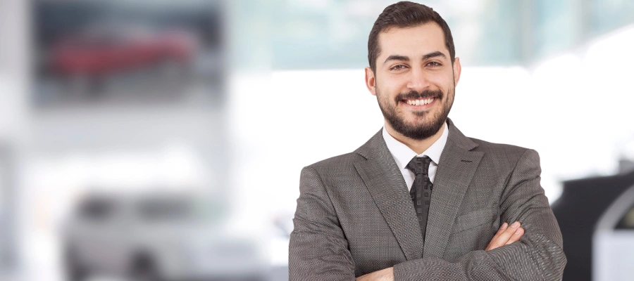 Businessman standing in automotive showroom, smiling with arms crossed.