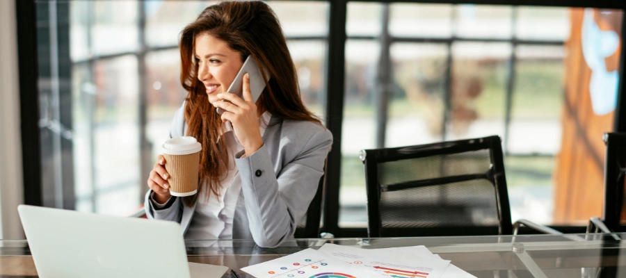 businesswoman sitting at office table with laptop and papers of statistics while talking on the phone and holding a cup of coffee